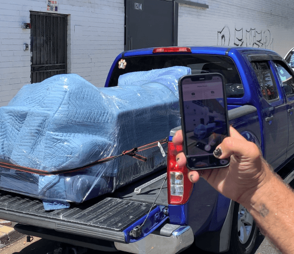 Close-up of a smartphone displaying a moving job in the Lugg app, with a wrapped couch loaded onto a blue truck in the background.