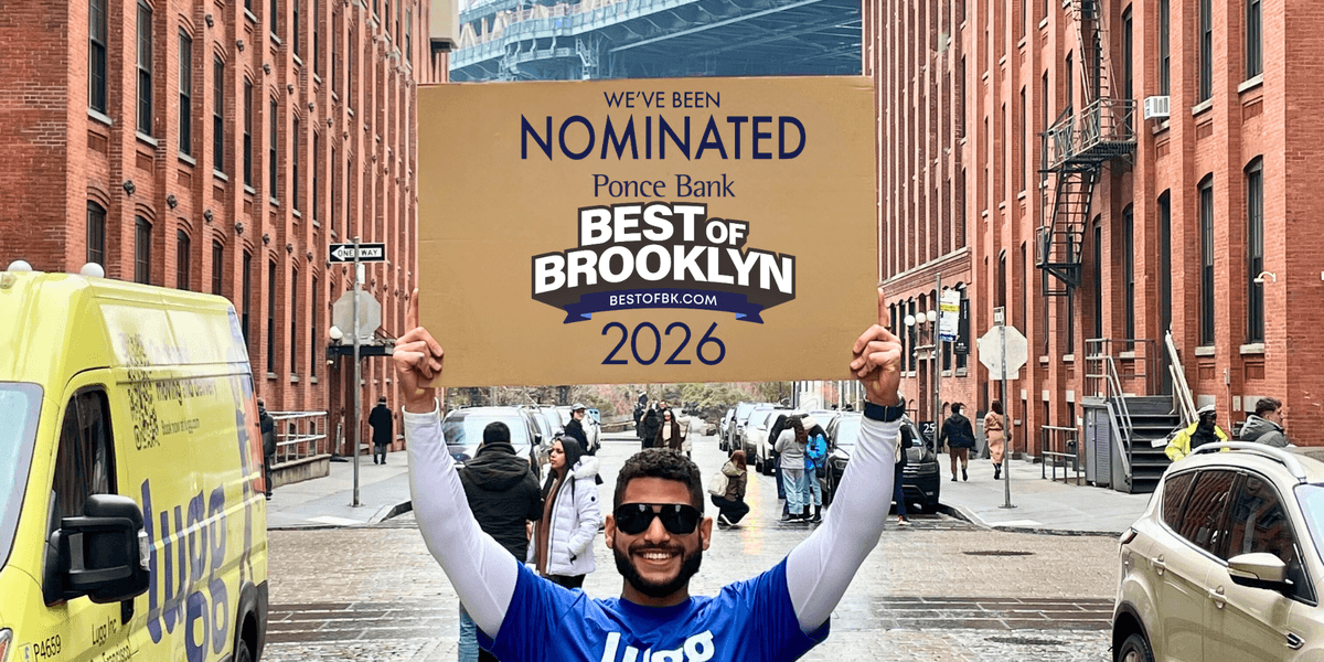 Lugg mover holding a Best of Brooklyn 2026 nomination sign on a street in Brooklyn with the Manhattan Bridge in the background.