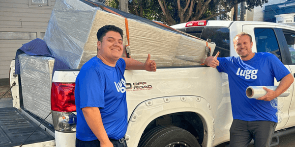 Two Lugg movers in branded blue shirts give a thumbs-up beside a white pickup truck, which holds a fully wrapped beige couch strapped in with moving pads and an orange tie-down.