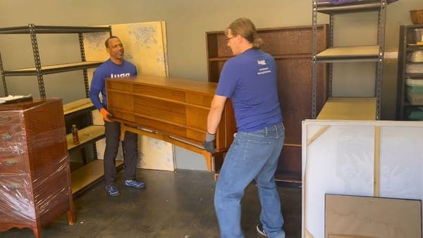 Two Lugg movers in branded gear carrying a mid-century wood dresser in a warehouse