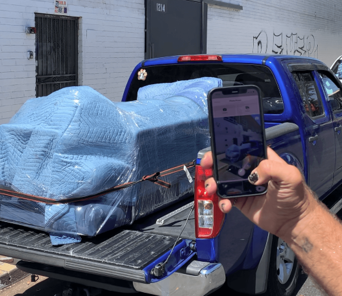 Close-up of a smartphone displaying a moving job in the Lugg app, with a wrapped couch loaded onto a blue truck in the background.
