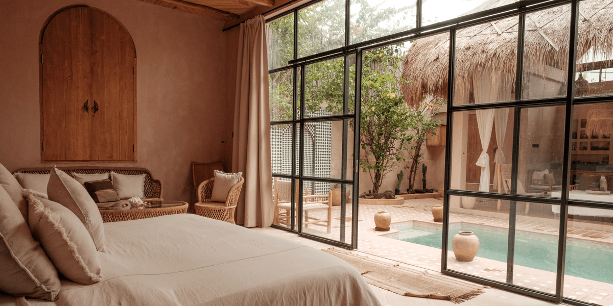 Neutral-toned bedroom with rattan chairs and a view of a private pool surrounded by tropical greenery through black-framed glass doors.