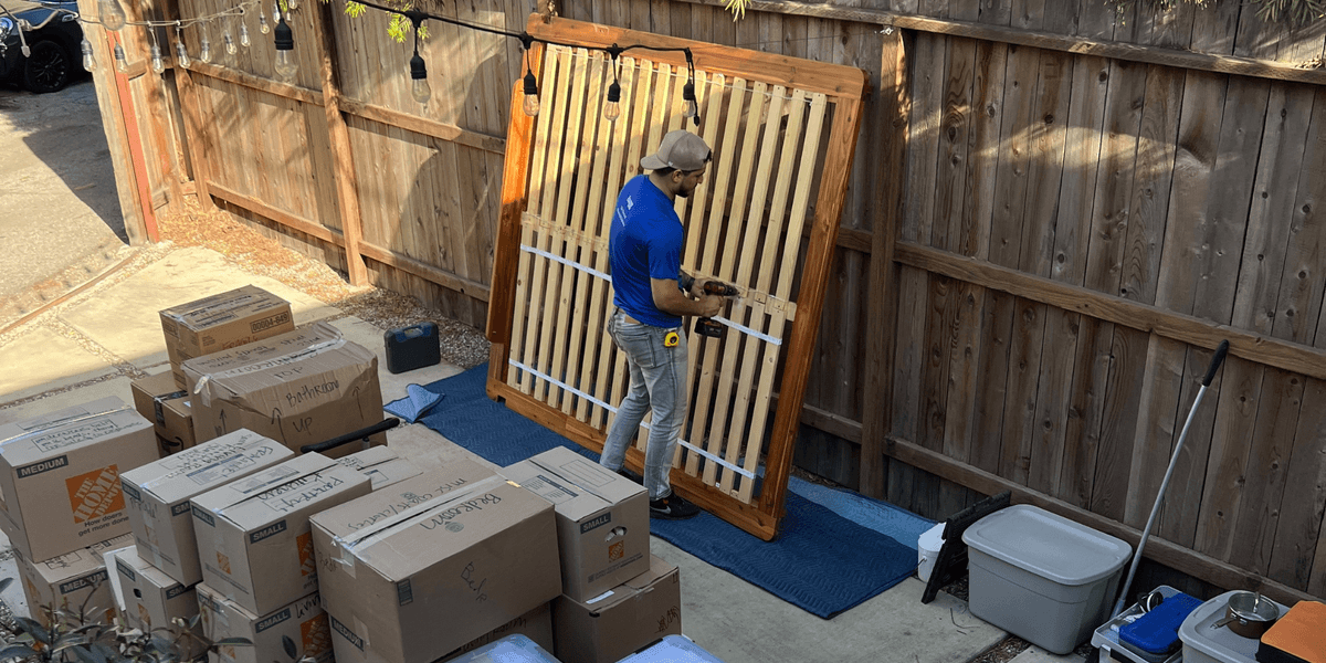 Lugg mover in blue shirt disassembling a wooden bed frame in a sunny backyard surrounded by packed boxes during a professional apartment move.