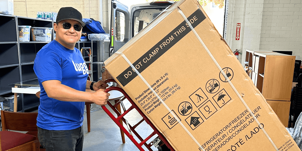 Lugg delivery professional smiling while using a dolly to move a boxed appliance for same-day delivery.