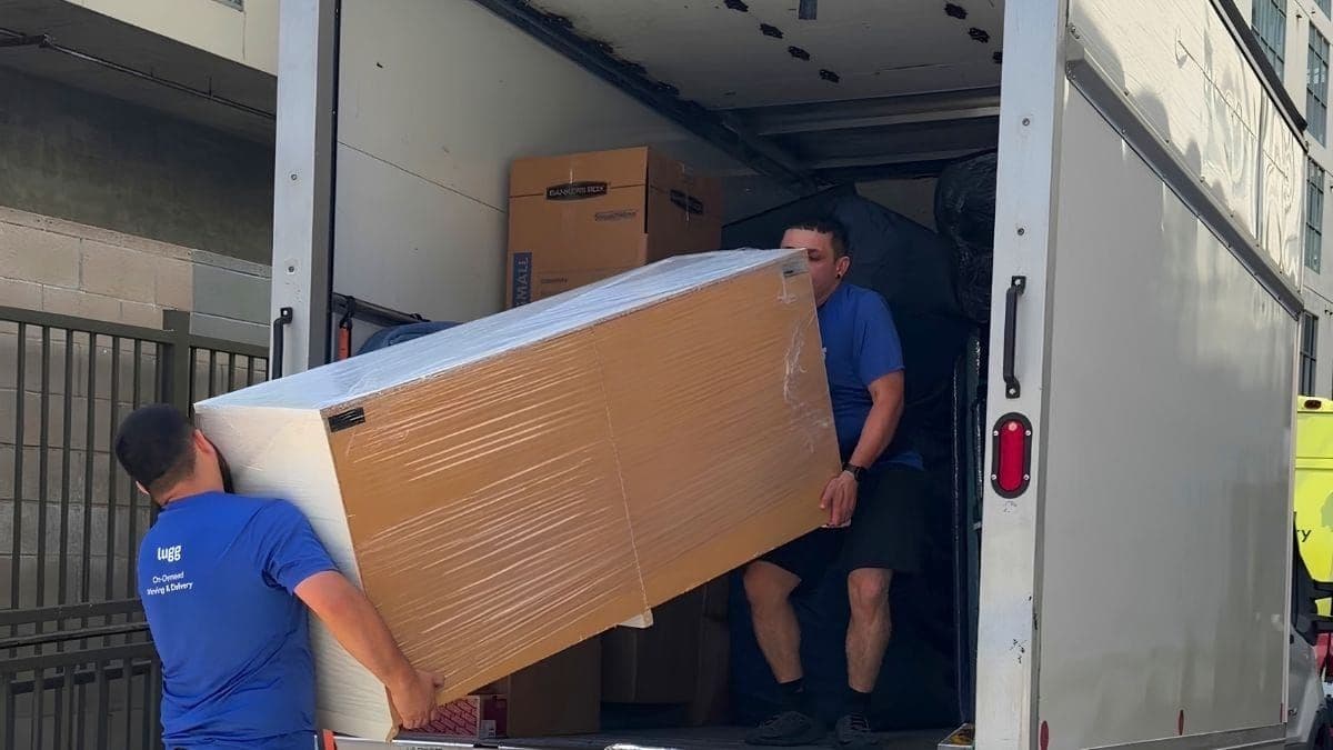 Two Lugg movers in blue shirts load a shrink-wrapped furniture piece into a box truck during a local move.
