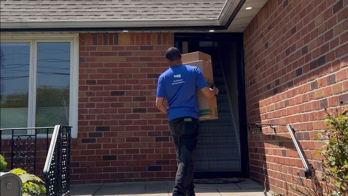 Lugg mover in a branded shirt carrying stacked moving boxes up to the front door of a brick home