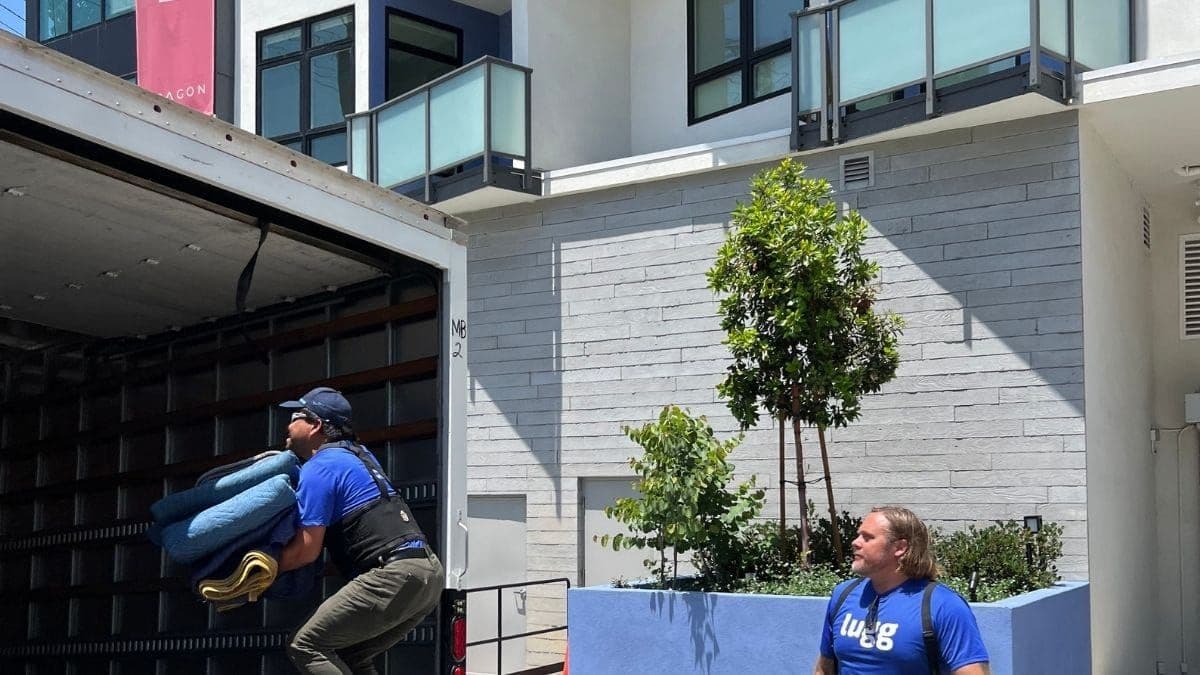 Two Lugg movers loading wrapped furniture into a box truck outside a modern multifamily apartment building with balconies