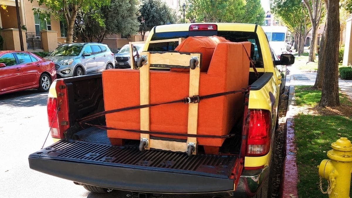 An orange secondhand couch strapped upright in a pickup truck bed on a tree-lined residential street — a cheap furniture find on its way home.