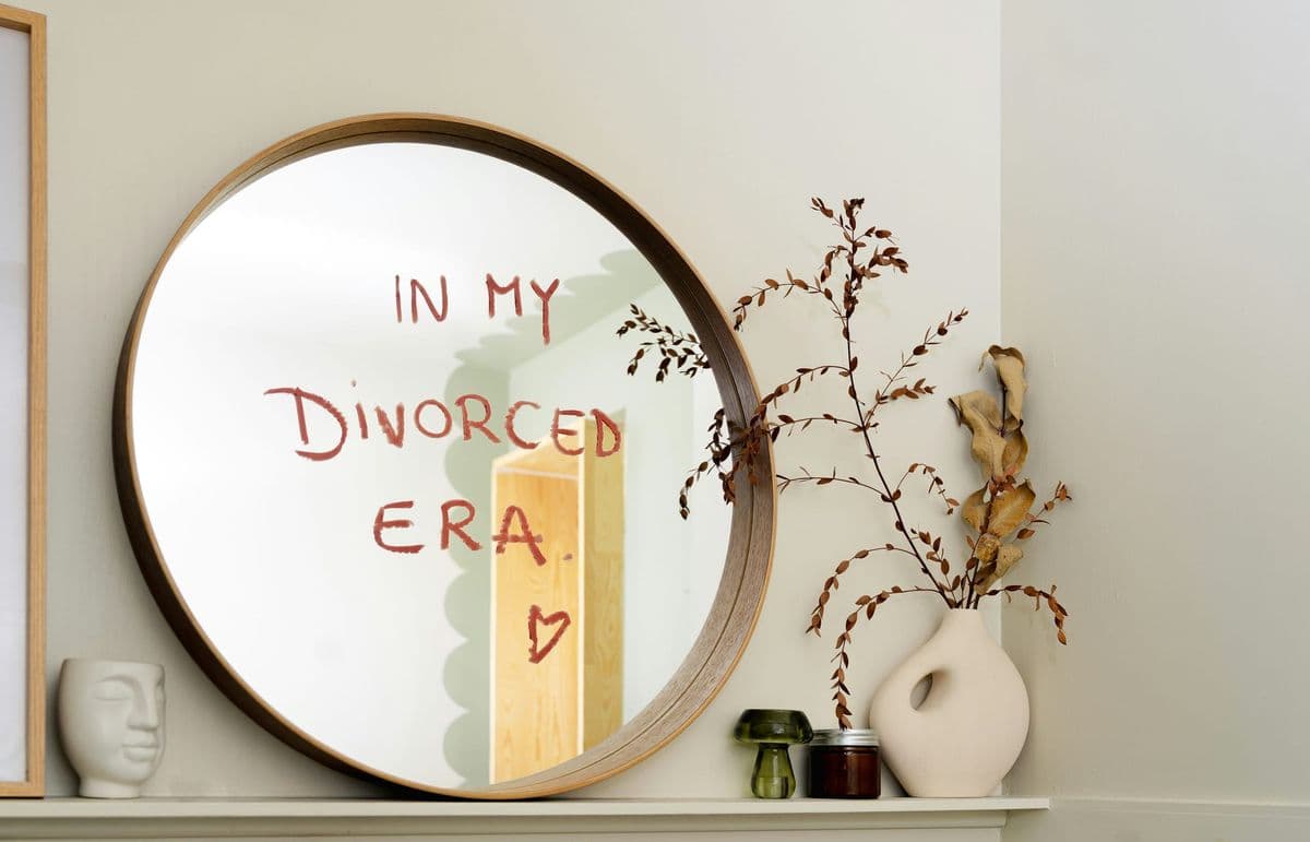 Round wood-framed mirror on a mantel with "In my divorced era" written in lipstick, styled with dried branches in a bright, freshly decorated room