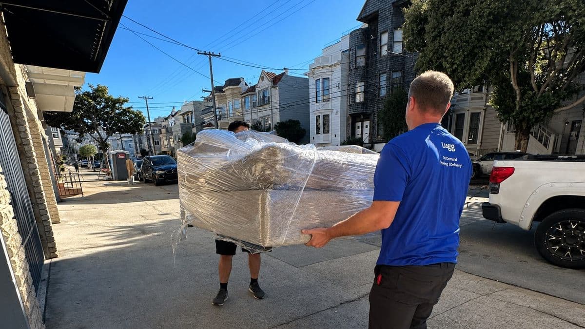 Two movers carry a shrink-wrapped couch up a San Francisco street, a pickup truck parked at the curb.