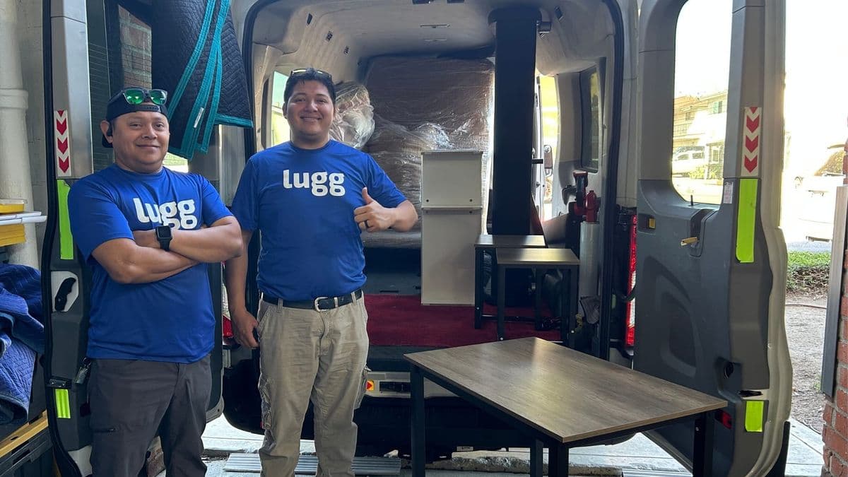 Two Lugg movers standing at the open rear of a loaded cargo van with a cabinet, a wood table and wrapped furniture from several stops