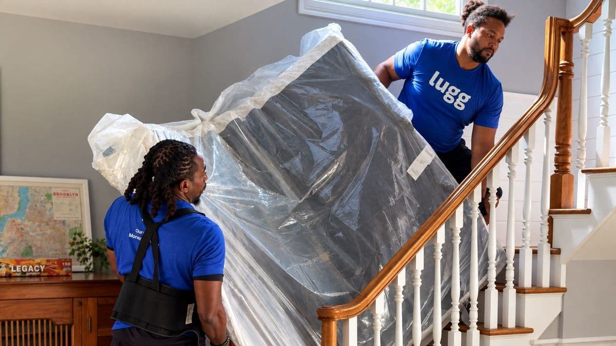 Two movers carry a wrapped mattress down a staircase inside a home.