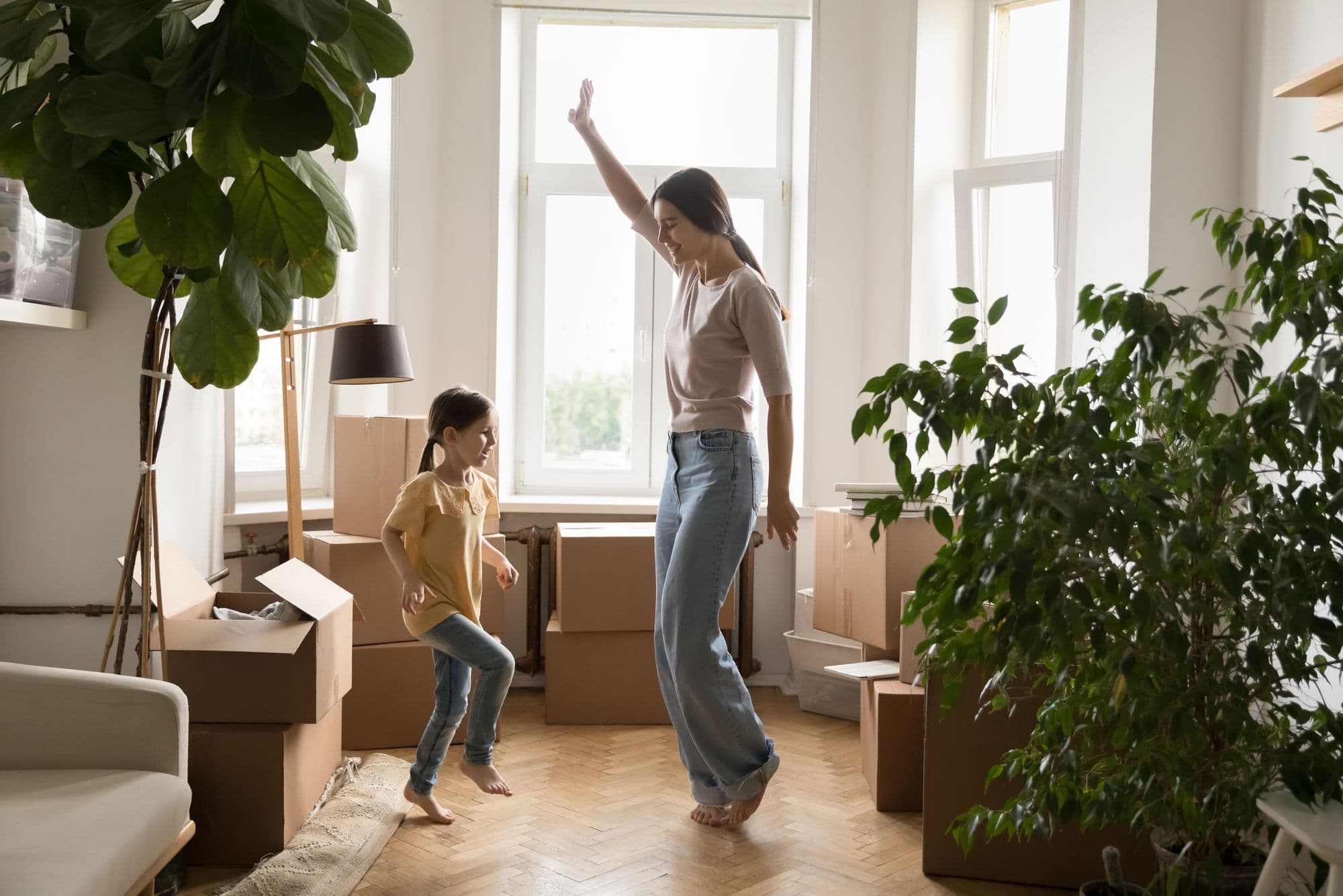 A mother and daughter dancing barefoot among moving boxes and houseplants in a bright, sunlit room during an eco-friendly move