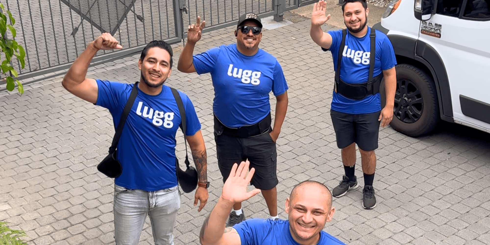 Four smiling Lugg movers in branded shirts waving outside a moving truck, showcasing friendly and on-demand moving help.