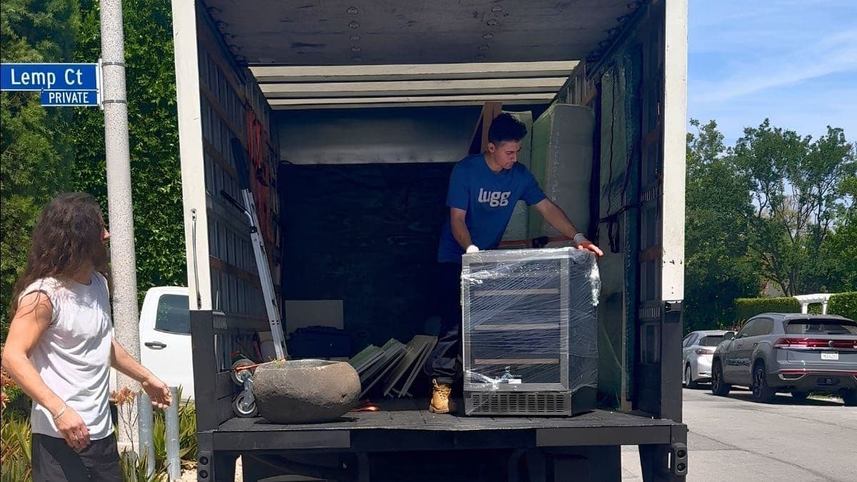 A Lugg driver loads a shrink-wrapped wine fridge into a box truck on the liftgate, with a hand truck and moving straps nearby.