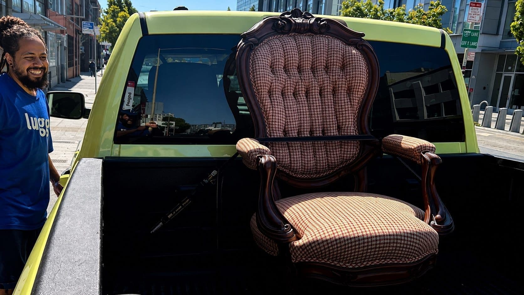 Lugg mover smiling beside an antique tufted armchair strapped upright in the bed of a Lugg pickup on a city street