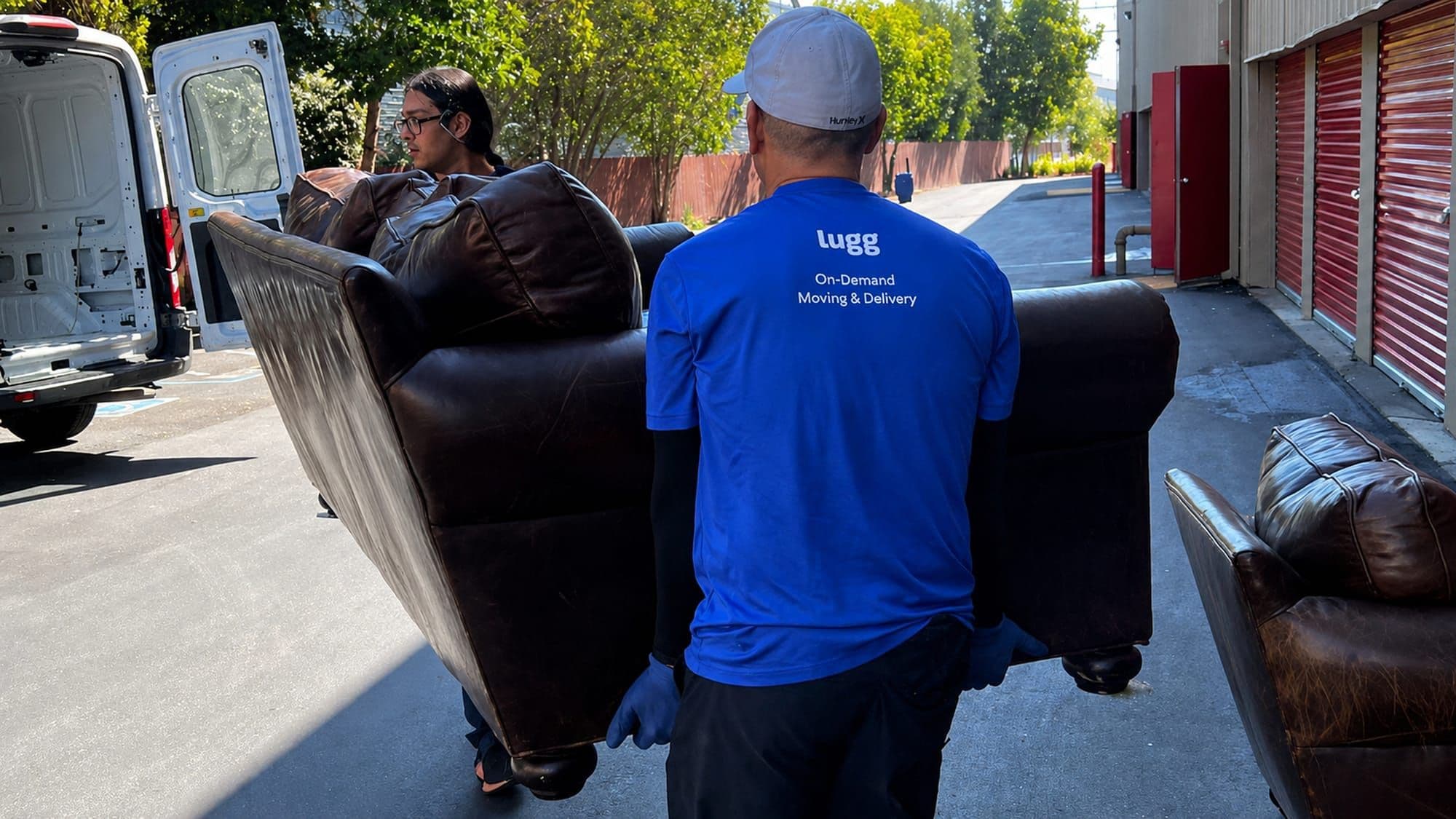 Two Lugg movers carrying a brown leather couch past a cargo van, one wearing a blue Lugg On-Demand Moving & Delivery shirt.