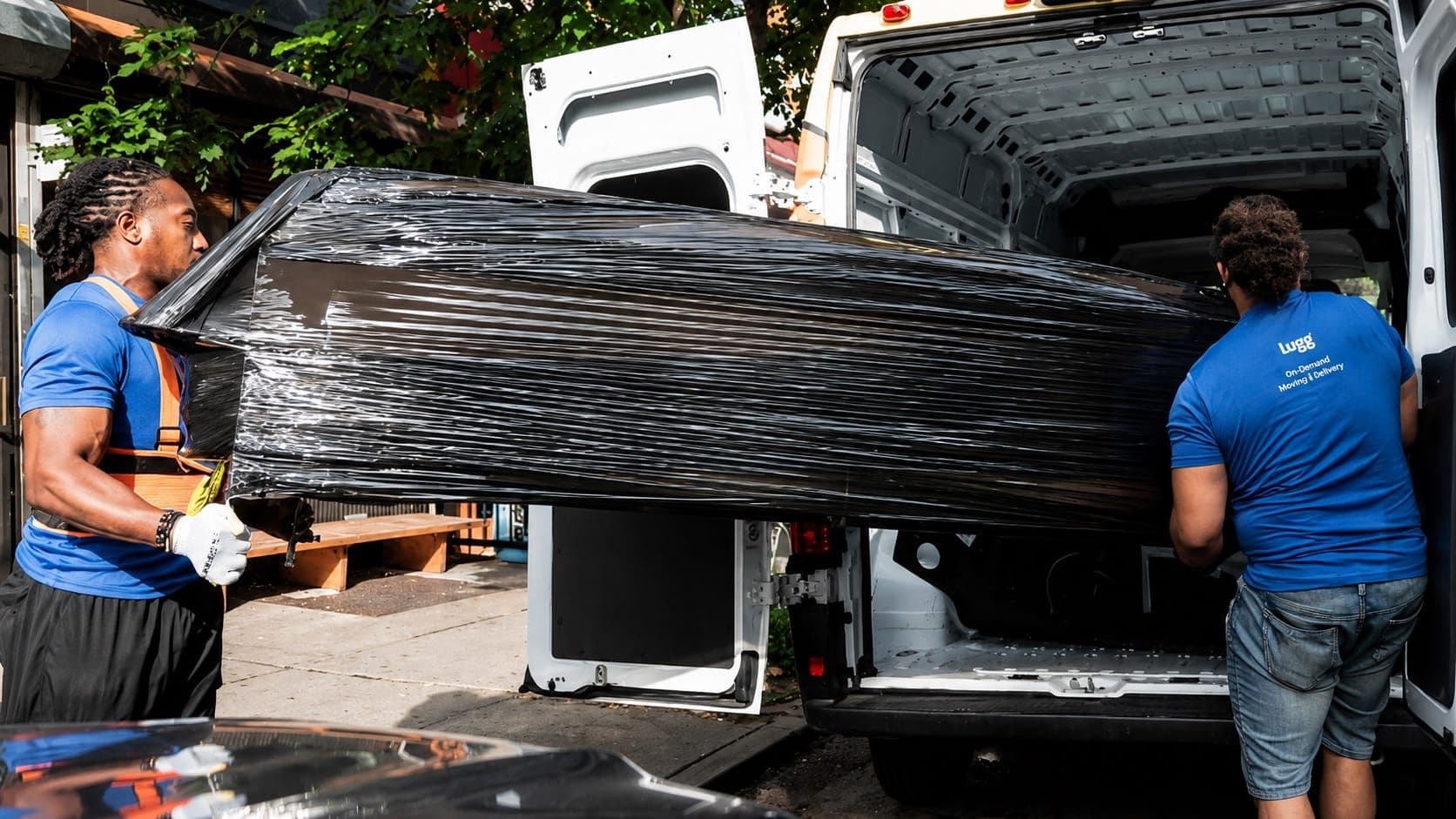 Two movers in blue shirts lift a large piece of shrink-wrapped furniture into the back of a white van on a city street.