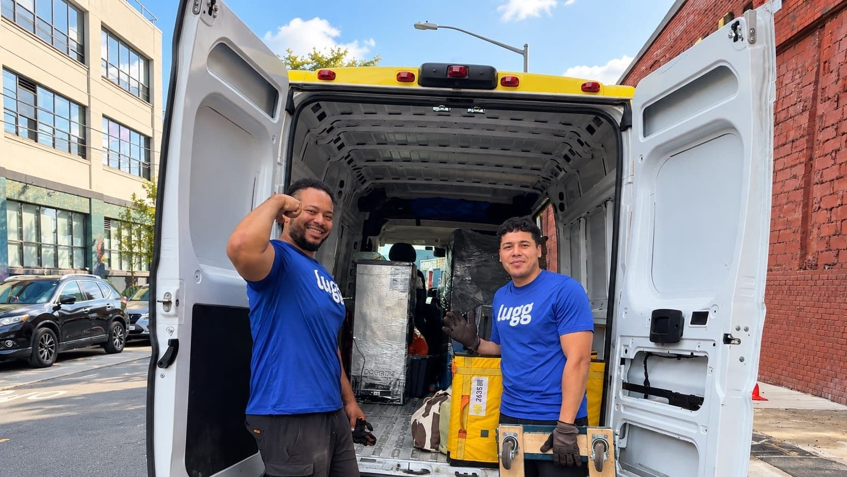 Two Luggers stand beside an open cargo van with moving equipment on a city street on a weekend morning.